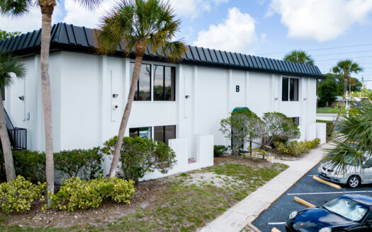 Two-story white commercial property with black roof and windows, surrounded by palm trees and shrubs, with parked cars in a partially visible parking lot.