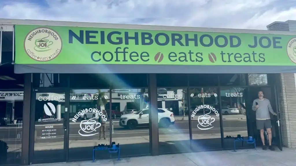 A person stands outside Neighborhood Joe, a commercial property coffee shop with a green sign reading "coffee eats treats." Cars are parked on the street in front.