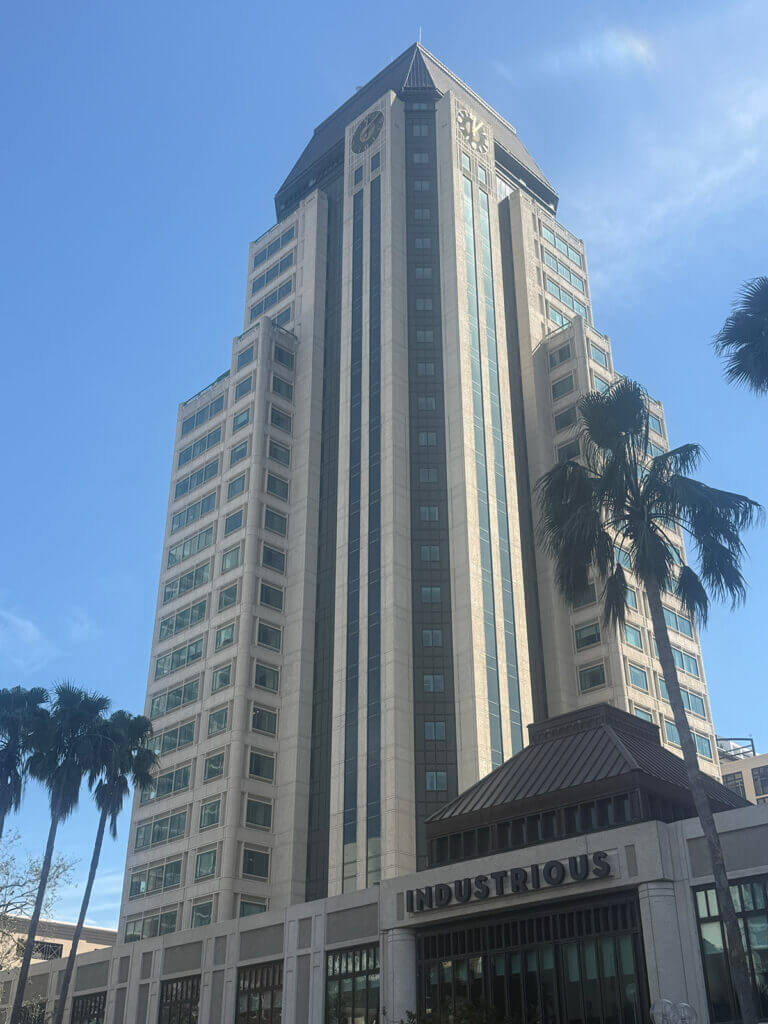 Tall modern commercial property with glass windows, palm trees in front, and a sign reading "INDUSTRIOUS" at the ground level, set against a clear blue sky.