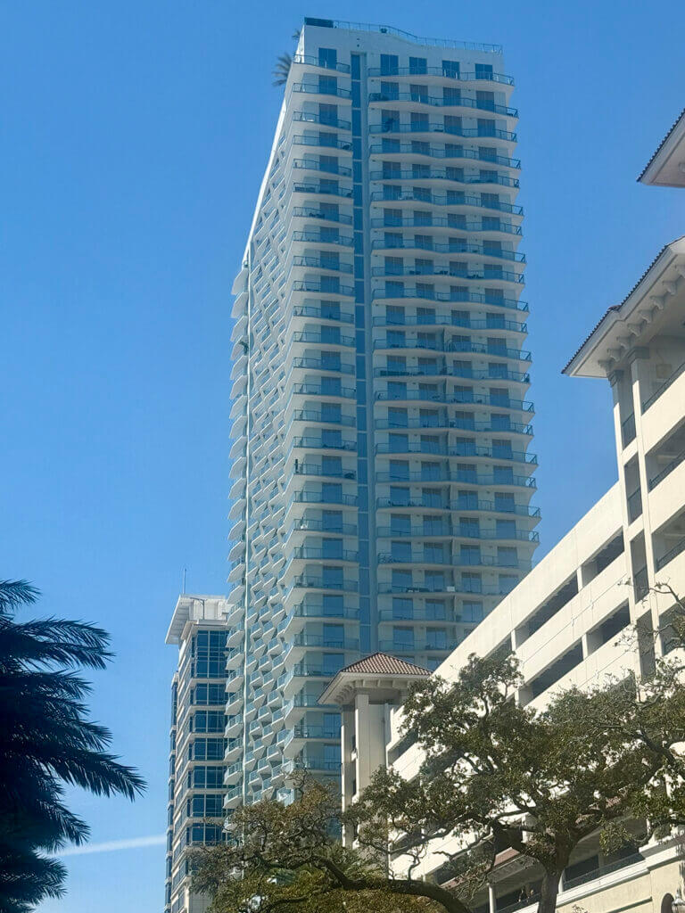 A tall modern glass high-rise residential building stands next to a multi-level parking garage and a nearby commercial real estate property on a clear, sunny day. Trees are visible in the foreground.