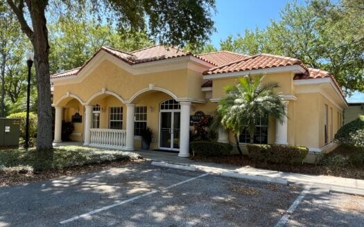 Charming single-story yellow stucco building at 11910 Boyette Rd in Riverview FL, featuring arched windows, white columns, and a red tile roof, nestled among trees with an empty parking lot.