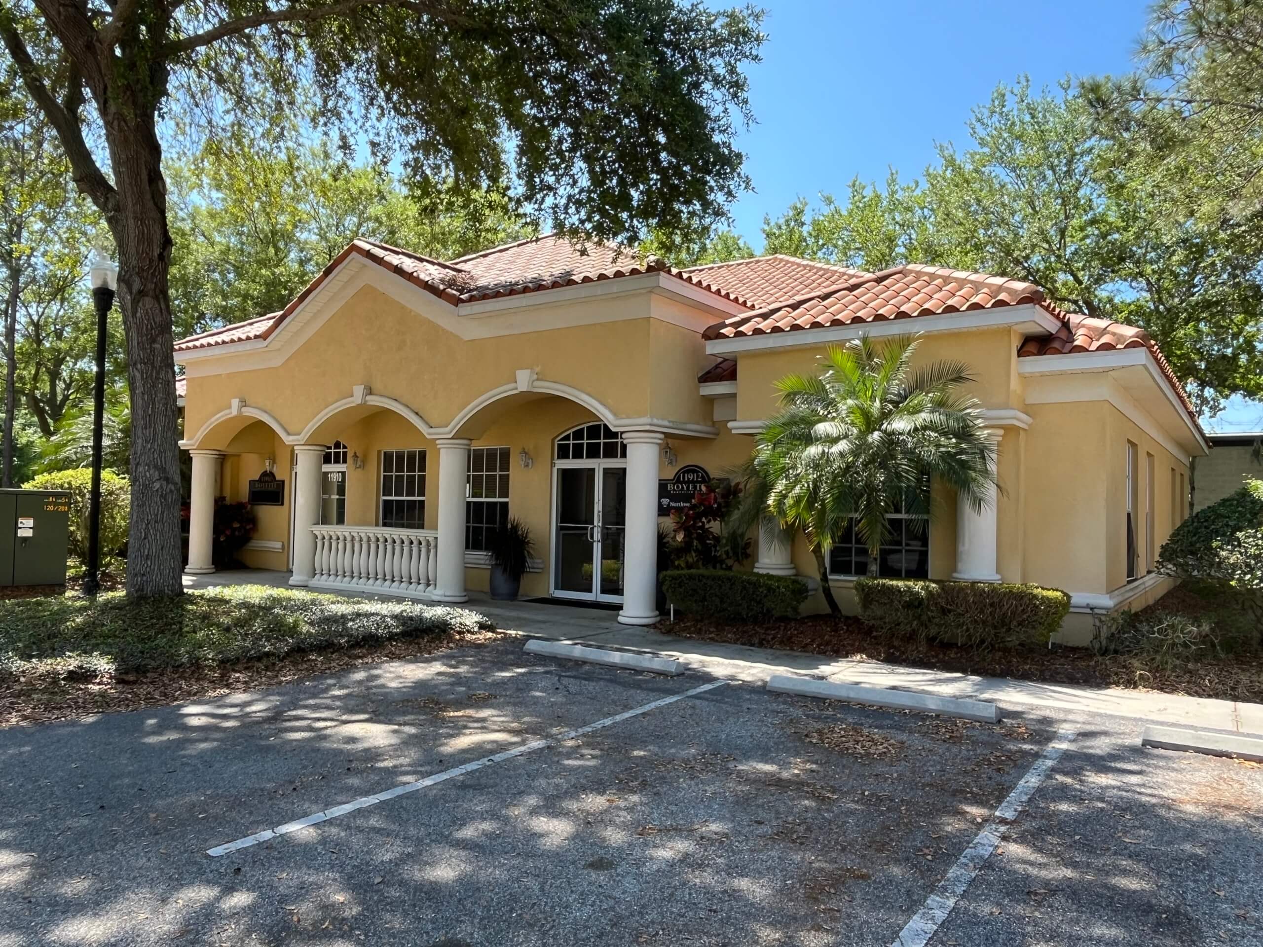 Charming single-story yellow stucco building at 11910 Boyette Rd in Riverview FL, featuring arched windows, white columns, and a red tile roof, nestled among trees with an empty parking lot.