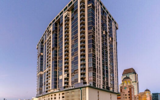 A tall, modern high-rise residential building with many windows stands on the corner of 1 Beach Drive SE in St. Petersburg FL, surrounded by trees and neighboring city buildings. Several units are currently leased.