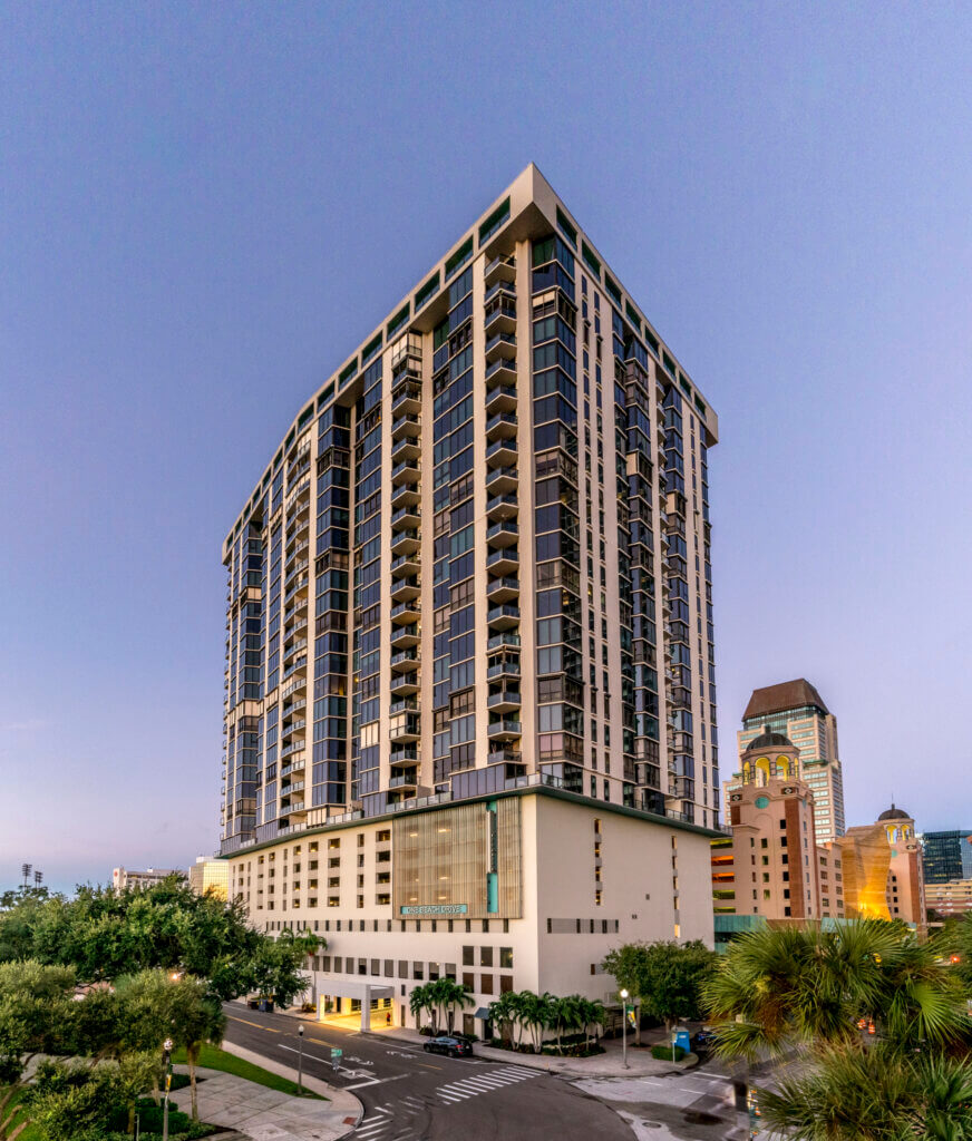 A tall, modern high-rise residential building with many windows stands on the corner of 1 Beach Drive SE in St. Petersburg FL, surrounded by trees and neighboring city buildings. Several units are currently leased.