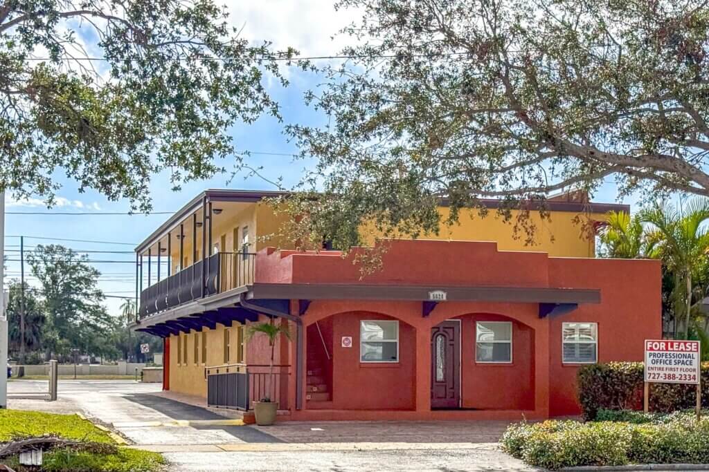 Two-story building with red and yellow exterior on Central Ave St. Petersburg features a covered walkway and "For Lease" sign for professional office space. Trees and small plants surround this leased commercial property in St. Petersburg FL.