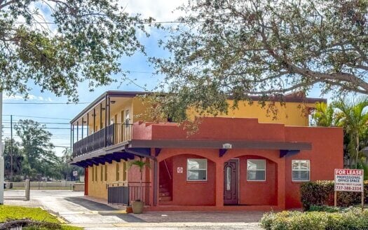 Two-story building with red and yellow exterior on Central Ave St. Petersburg features a covered walkway and "For Lease" sign for professional office space. Trees and small plants surround this leased commercial property in St. Petersburg FL.