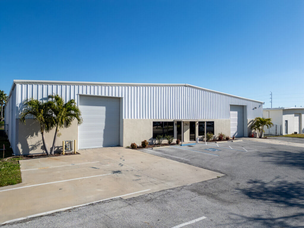 A one-story industrial warehouse with white metal siding, two large garage doors, glass entry doors, and palm trees stands at 2963 44th Ave. N., St. Petersburg, FL, with an empty parking lot under a clear blue sky.