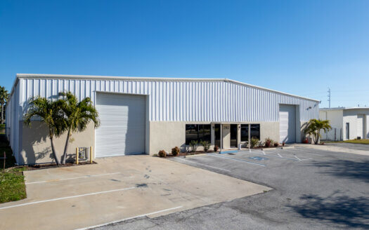 A one-story industrial warehouse with white metal siding, two large garage doors, glass entry doors, and palm trees stands at 2963 44th Ave. N., St. Petersburg, FL, with an empty parking lot under a clear blue sky.