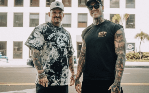 Two tattooed men stand on a street in front of One Beach Drive, both wearing casual summer clothes, sunglasses, and hats—a scene capturing the laid-back vibe of St. Pete Rising near Bayfront Tower Retail.