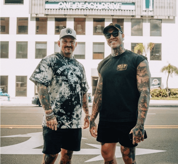 Two tattooed men stand on a street in front of One Beach Drive, both wearing casual summer clothes, sunglasses, and hats—a scene capturing the laid-back vibe of St. Pete Rising near Bayfront Tower Retail.