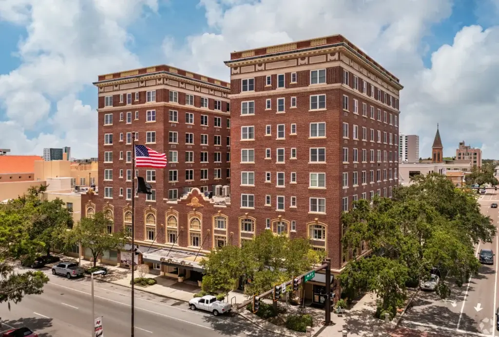A historic, multi-story brick hotel building on First Avenue in St. Petersburg, with an American flag in front, set along a tree-lined street in an urban area under a partly cloudy sky.