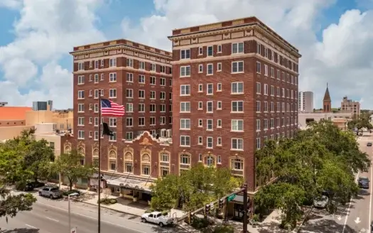 A historic, multi-story brick hotel building on First Avenue in St. Petersburg, with an American flag in front, set along a tree-lined street in an urban area under a partly cloudy sky.