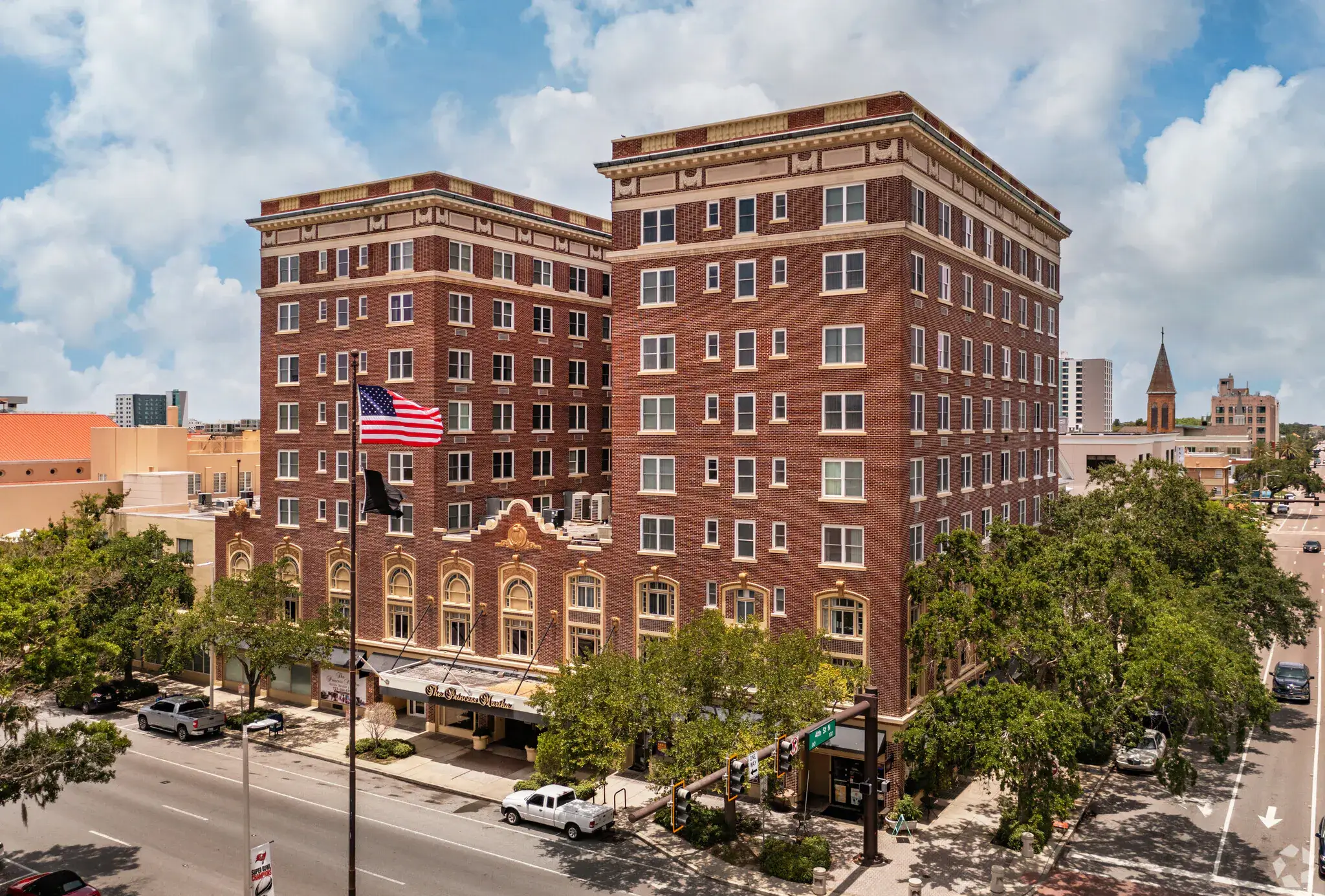 A historic, multi-story brick hotel building on First Avenue in St. Petersburg, with an American flag in front, set along a tree-lined street in an urban area under a partly cloudy sky.