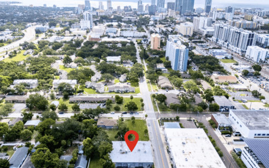 Aerial view of a cityscape in St. Petersburg with a red location marker over a building in the foreground; high-rise buildings and 14th St, FL 33705 visible in the background.