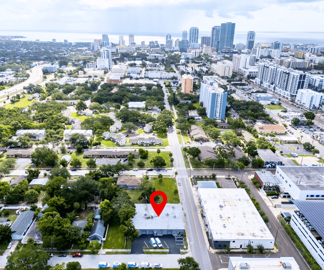 Aerial view of a cityscape in St. Petersburg with a red location marker over a building in the foreground; high-rise buildings and 14th St, FL 33705 visible in the background.