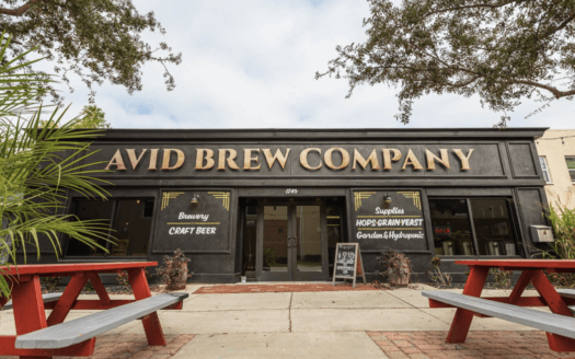 A black storefront with gold lettering reading "Avid Brew Company"—the Avid Brewing Property—for sale, flanked by windows advertising craft beer and brewing supplies, with red picnic tables in front. As seen in St. Pete Catalyst.