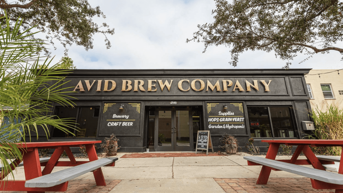 A black storefront with gold lettering reading "Avid Brew Company"—the Avid Brewing Property—for sale, flanked by windows advertising craft beer and brewing supplies, with red picnic tables in front. As seen in St. Pete Catalyst.