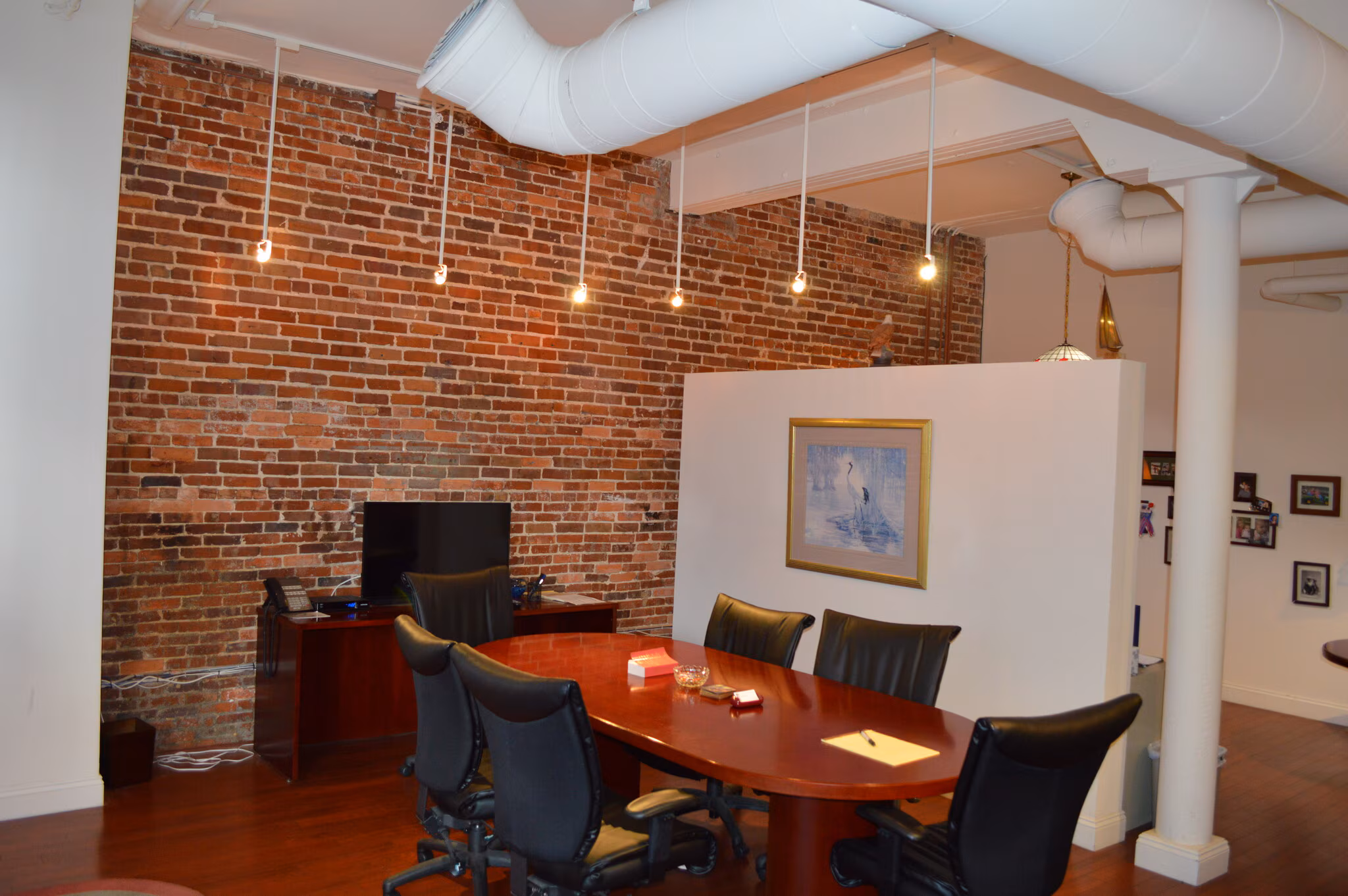 A modern office space with exposed brick walls, a wooden conference table with chairs, a desk with a monitor, and hanging ceiling lights.