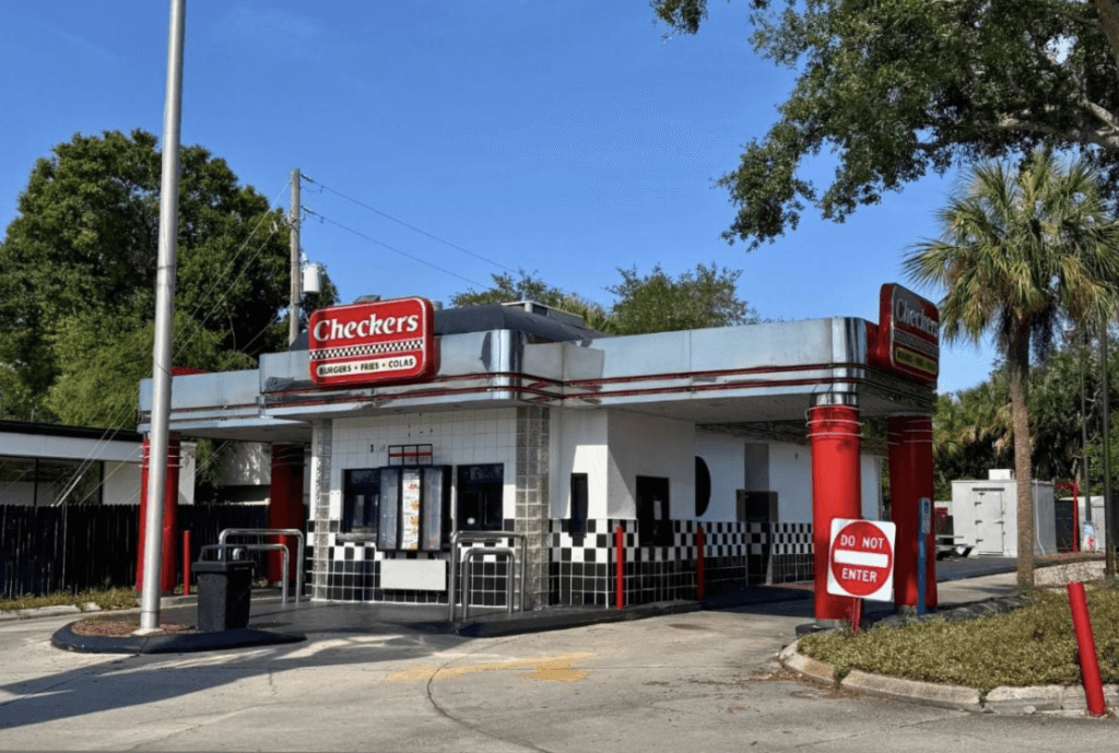 A Checkers fast-food restaurant, leased at 3501 34th St. N, St. Petersburg, features a retro design with black and white checkered tiles, red accents, and a drive-thru lane under a clear blue sky.